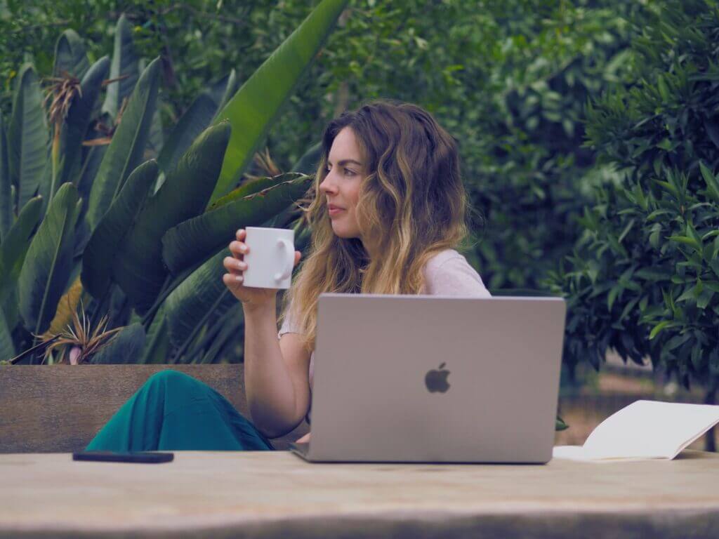 Spirituelle Webdesignerin Matthea Adrienne sitzt vor einer grünen Wand aus Bananenstauden, vor ihr ein aufgeklappter Laptop, in ihrer Hand eine Kaffeetasse und schaut gedankenverloren in die Ferne links aus dem Bild
